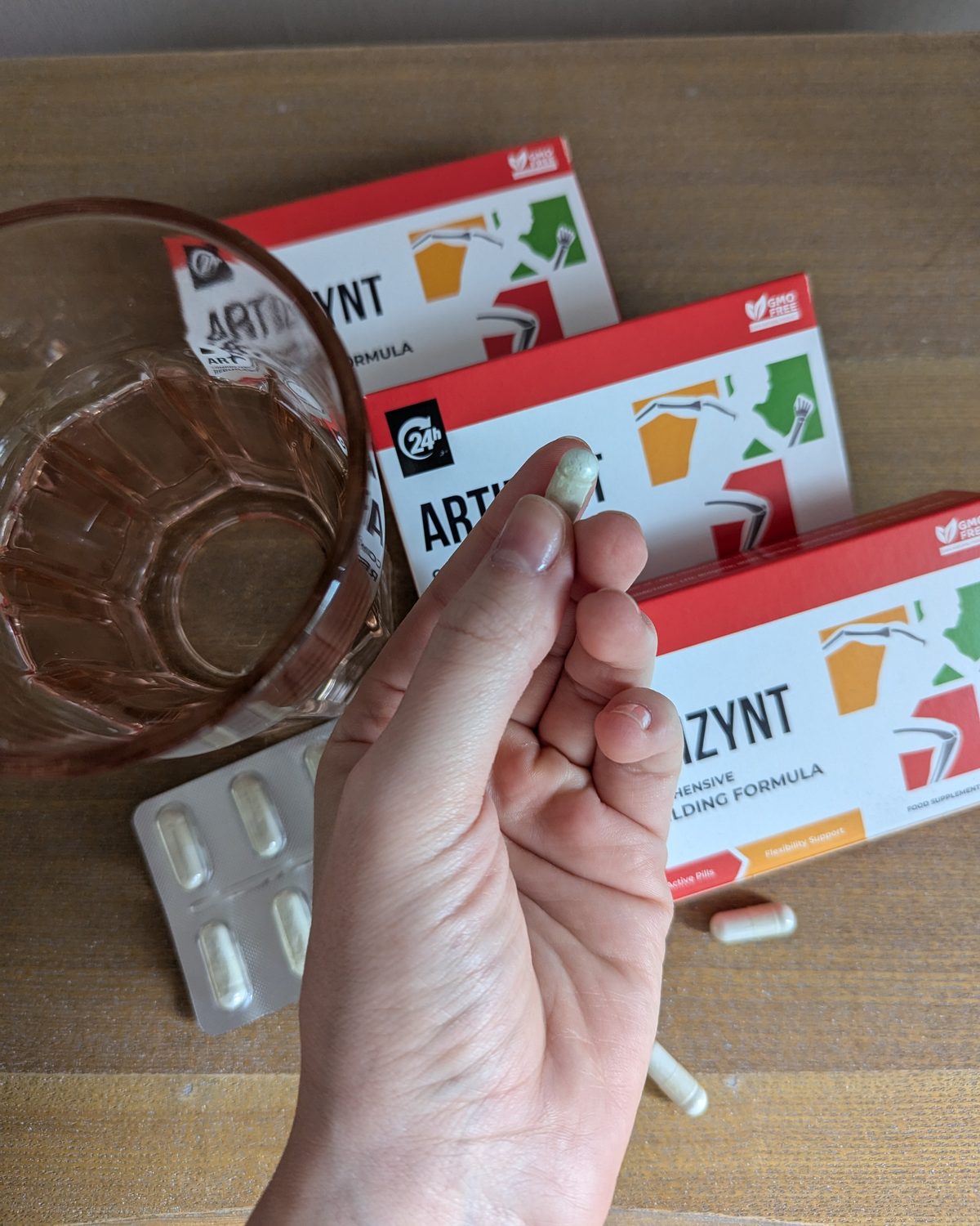 Eine weiße ArtiZynt-Kapsel zwischen zwei Fingern, dahinter drei Packungen, ein Glas Wasser und ein Blister auf einem Holztisch